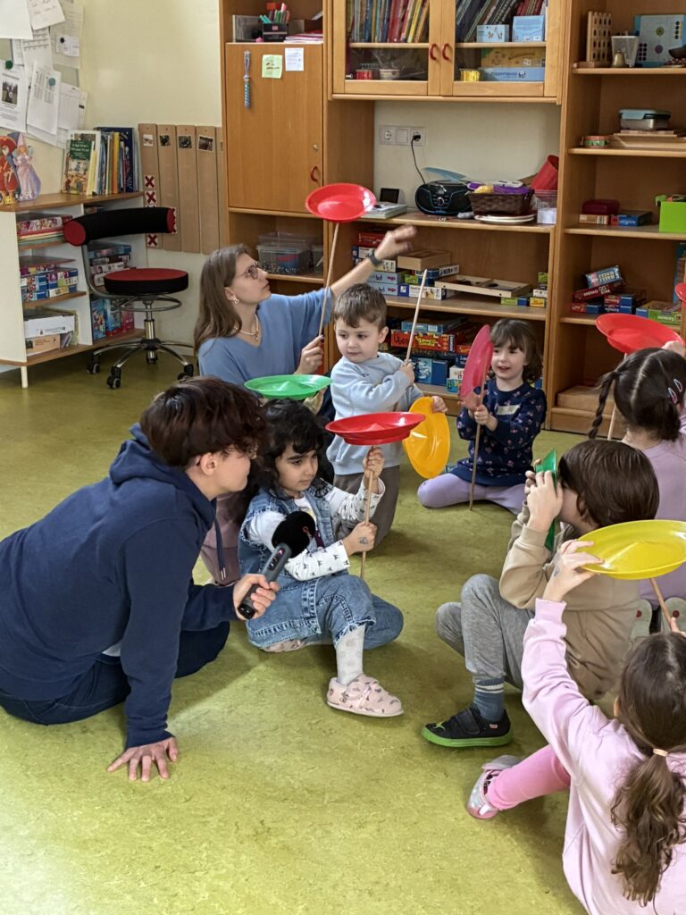 Die Kinder üben das Bewegen eines Tellers auf der Spitze eines Stabs, im Vordergrund hält die Reporterin einem Mädchen das Mikrofon hin.