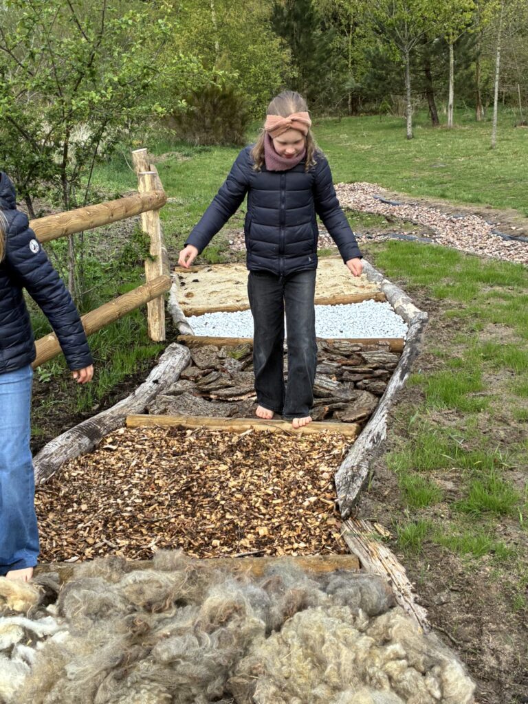 Ein Mädchen begeht den Barfußpfad, leicht zögernd aufgrund der unterschiedlichen Untergründe, wie Holzschnipsel. Vorn im Bild ist ein Bereich mit Schafswolle ausgelegt.