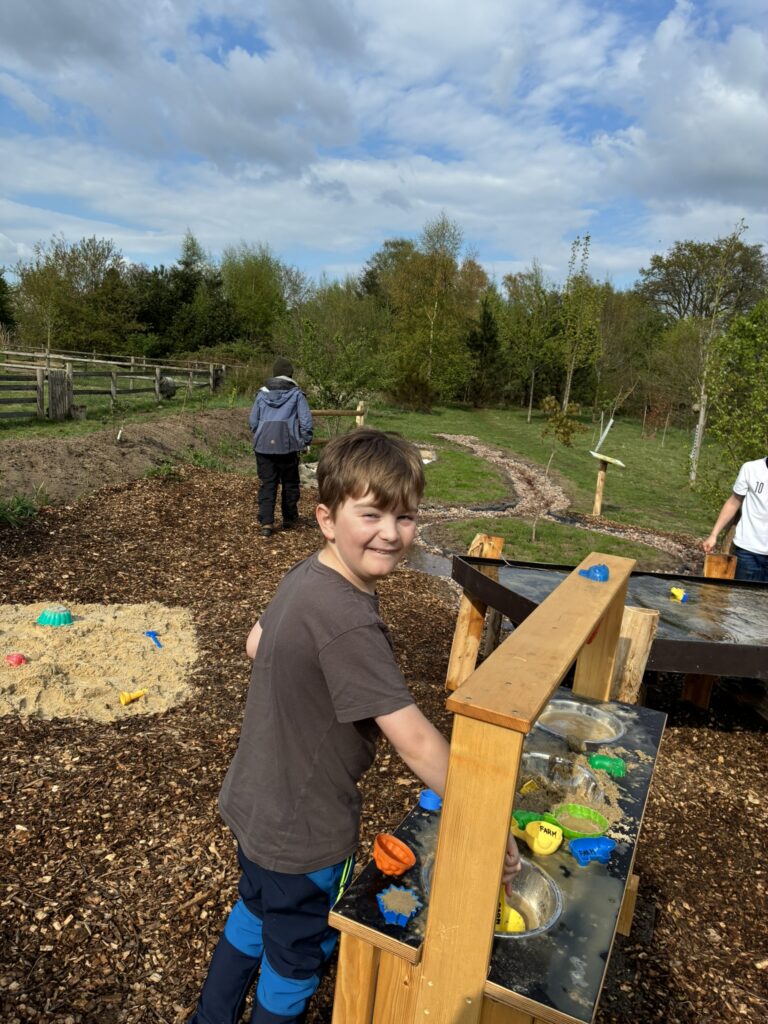 Ein Junge im Grundschulalter lächelt in die Kamera während er mit einer Schaufel Sand verrührt in der Matschküche.