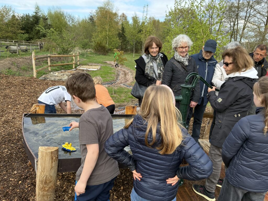 Vertreter:innen des Kiwanis Clubs Bremen Ginkgo erproben die Wasserpumpe. Kinder stehen um das Wasserbecken und sind spielend auf dem weiteren Gelände zu sehen.