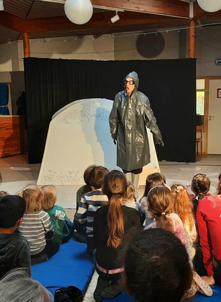 Beim Kindertheater verfolgen Kinder auf dem Boden sitzend, mit dem Rücken zum Betrachter einen Darsteller, gekleidet ganz in Grau, schwarzer Gesichtsmaske und mit weit aufgerissenem Mund und großen Augen.