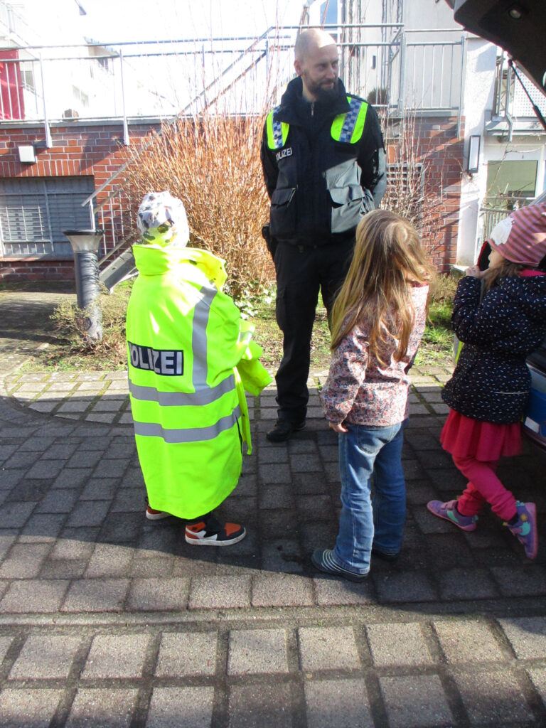 Drei Kitakinder stehen vor einem freundlich blickenden Polizisten und scheinen ihn zu befragen. Eins der Kinder trägt eine Leuchtjacke mit dem Aufdruck POLIZEI