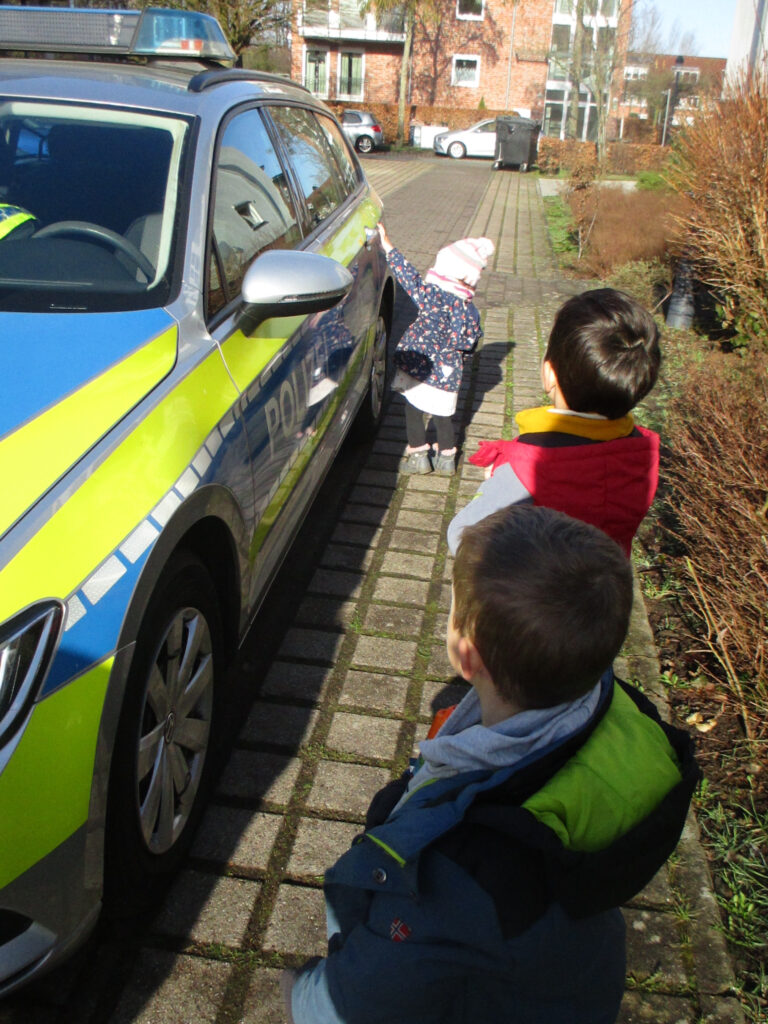 Drei Kita-Kinder schauen sich die Seite eines Polizeiwagens an. Ein Mädchen ist dabei, die hintere Tür zu öffnen.
