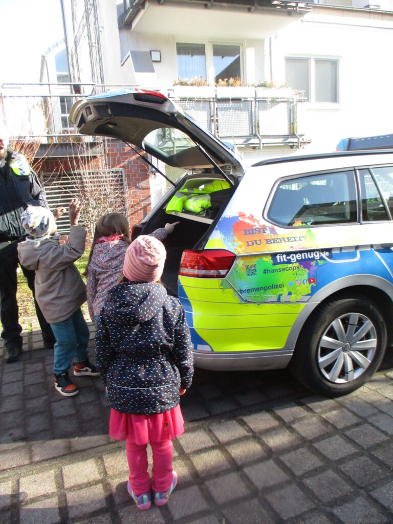 Drei Kita-Kinder erkunden gemeinsam das Polizeitauto. Sie schauen gemeinsam in den Kofferraum, auf den ein Mädchen zeigt.