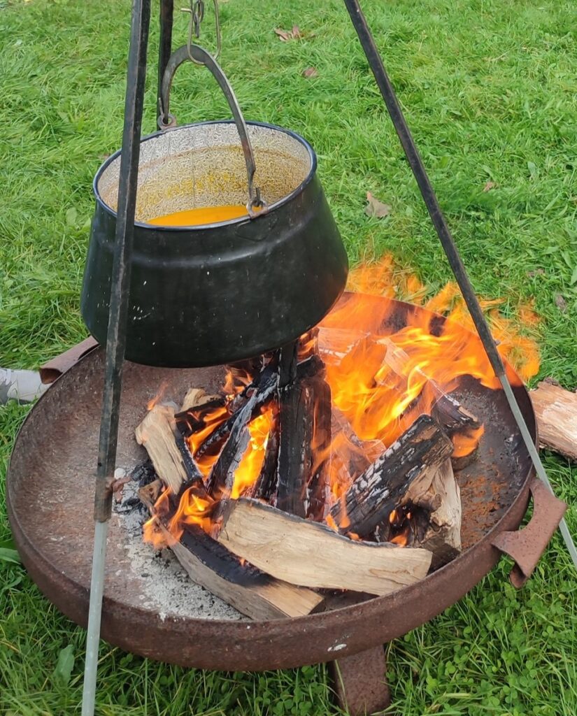Ein loderndes Lagerfeuer in einer Feuerschale, die auf grünem Rasen steht. Darüber hängt ein Kochtopf mit Kürbissuppe.