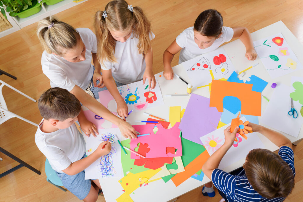 Mutter oder Lehrerin mit Kindern um einen Tisch mit Kreativmaterialien. Die Kinder malen, schneiden aus und sind kreativ.