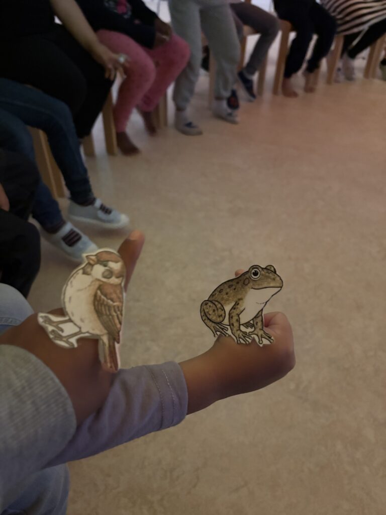 Auf Kinderhänden sind ein Frosch sowie ein Spatz aus Papier gelegt - zwei Figuren aus einem Buch mutmaßlich. Im Hintergrund sind die Beine der weiteren Kinder aus einem Sitzkreis zu erkennen.