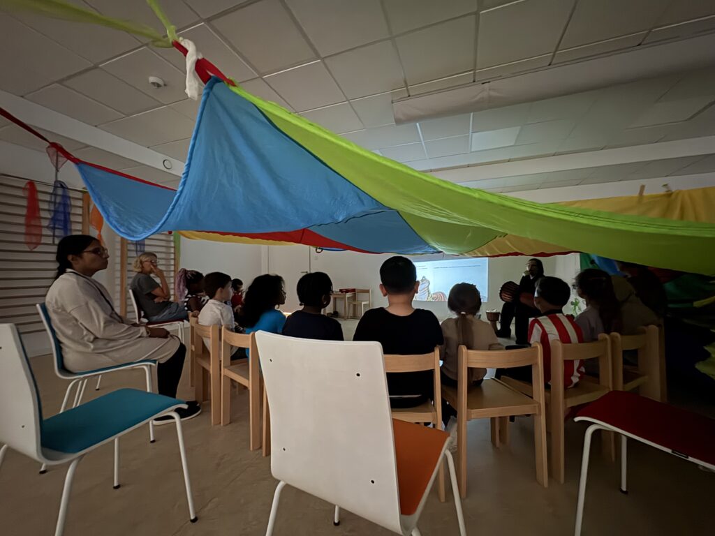 Kinder sitzen mit dem Rücken zum Betrachter im Halbkreis unter einem bunten Schwungtusch vor einem auf eine Wand projizierten Bild.