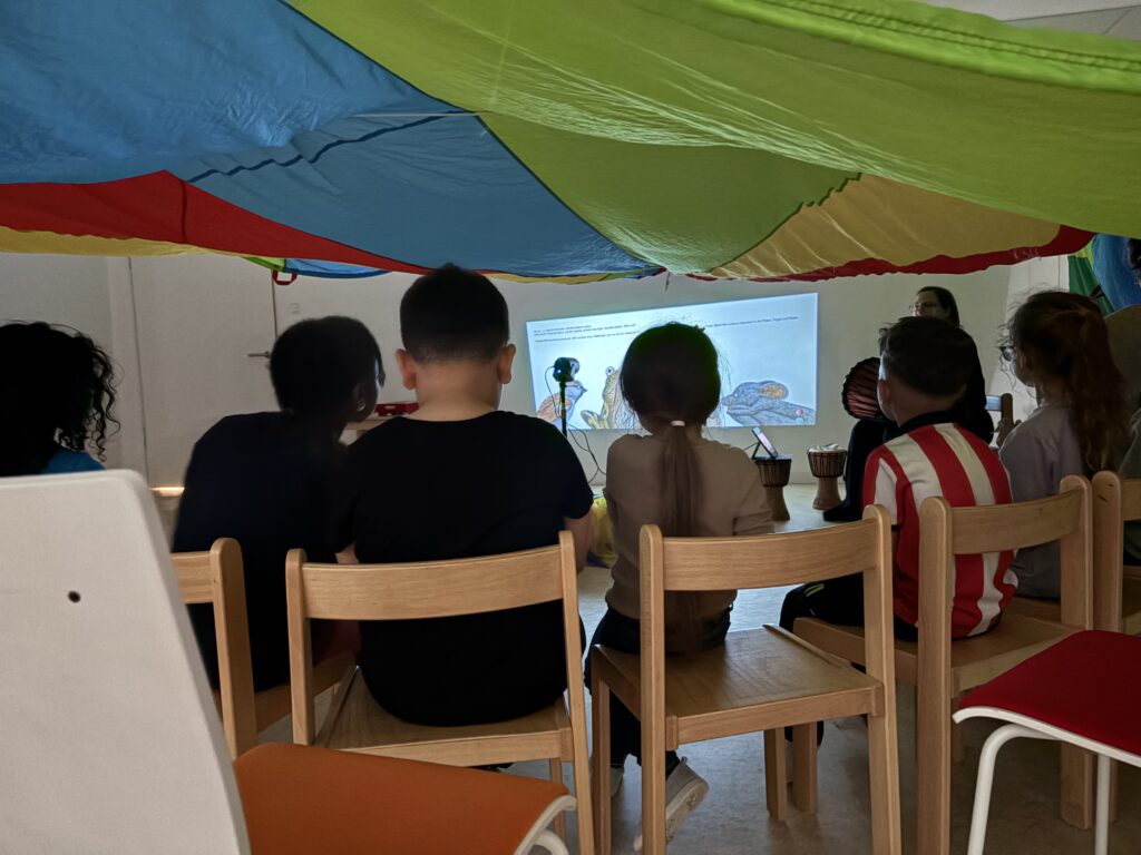 Kinder sitzen mit dem Rücken zum Betrachter im Halbkreis unter einem bunten Schwungtusch vor einem auf eine Wand projizierten Bild.